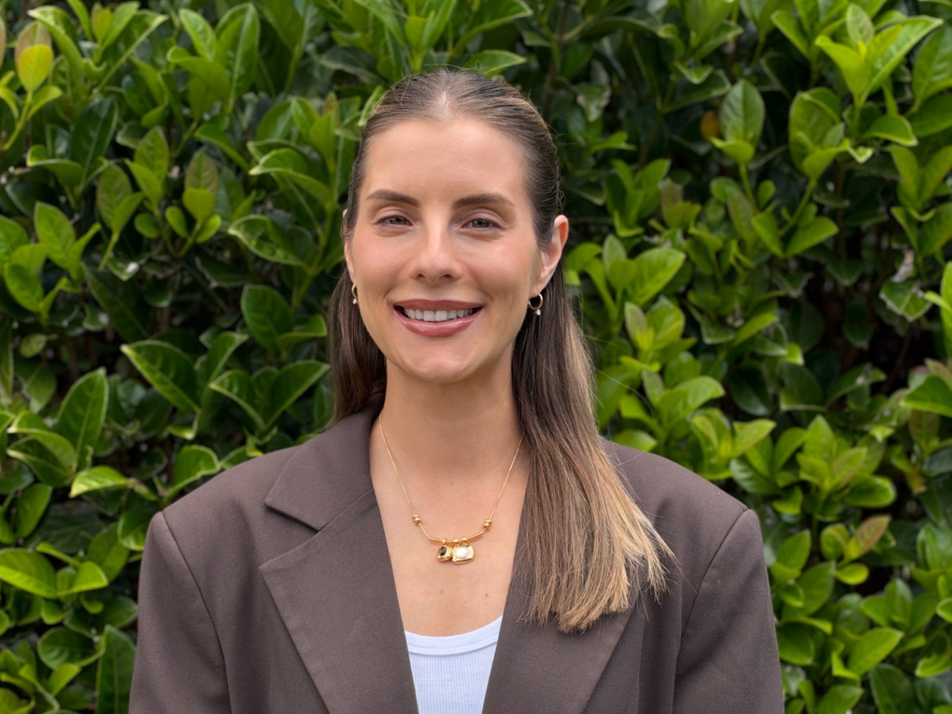 Nikki smiling in front of a green leafy hedge, wearing a brown blazer over a white top