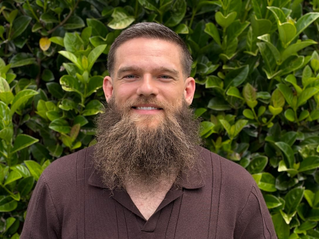 The founder of That Ability smiling in front of a green leafy hedge, wearing a dark brown knit polo shirt
